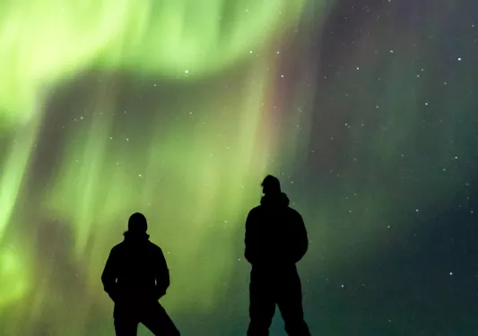Silhouettes of 2 people standing on a cliff with the Northern Lights in the sky