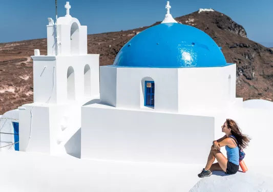 Woman sitting on the roof of a white, Greek house, with large cliffs in the background