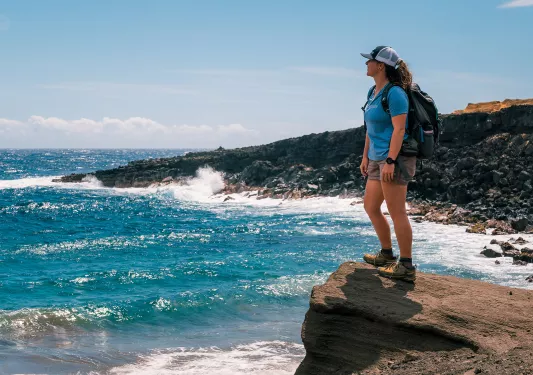 Woman wearing a backpack, standing on a cliff looking out to the ocean