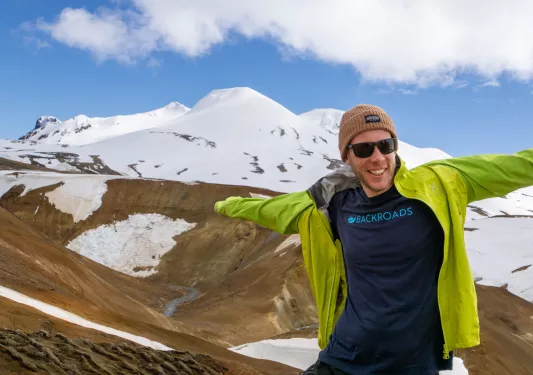 Man wearing a green jacket with his arms open, with snowy mountains in the background