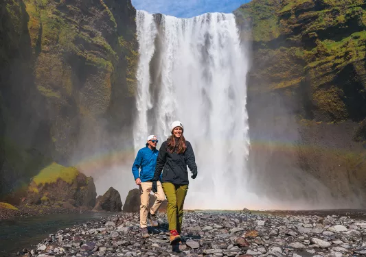 Man and woman walking on rocks in front of a large waterfall