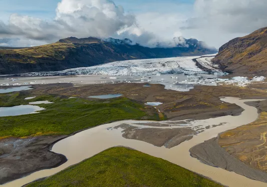 Valley with a brown river, in front of a large lake with ice caps behind it