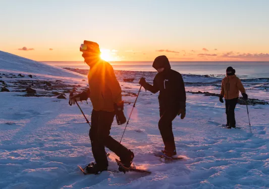 Three people wearing snow gear, walking on a field of snow by the ocean