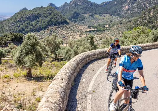 Two women riding a bike on a road with a large grass and tree valley in the background