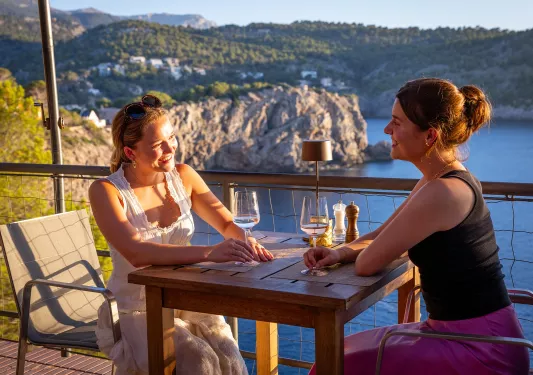 Two women sitting at a table on a patio, holding glasses of wine with a lake in the background