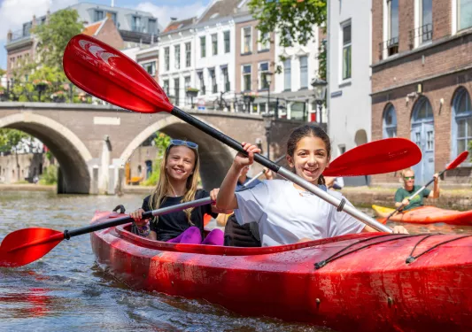 Two girls paddling in a red kayak on a river, with a town in the background