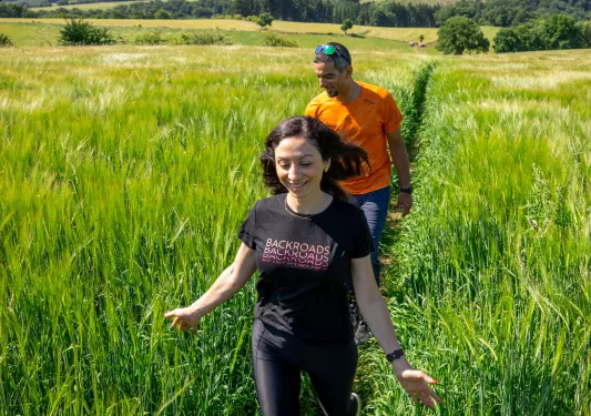 Man and woman walking through a field of tall weeds with large hills in the distance