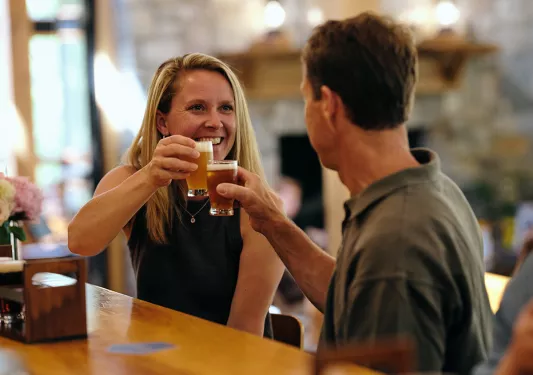 Man and woman sitting at a bar, smiling while raising glasses of beer