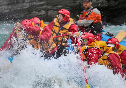 Group of people wearing red jackets and helmets, paddling on a raft in an active river