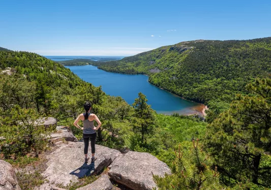 Woman standing on top of a boulder, looking out to a large lake and forest