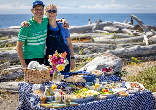 Man and woman smiling and hugging, standing in front of a picnic table full of snacks