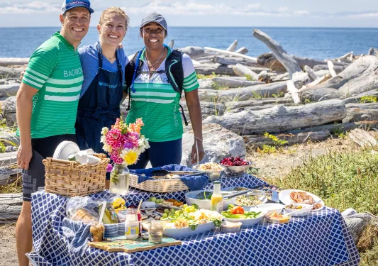 One man and two women standing behind a table full of snacks, with the ocean in the background