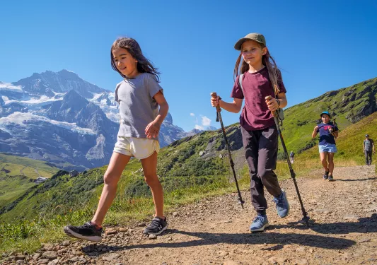 Two girls with hiking poles, walking on a gravel trail with large mountains in the distance