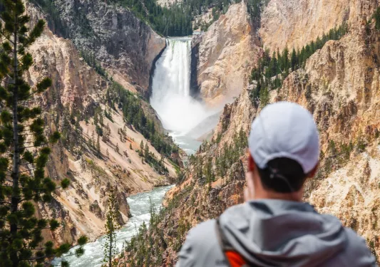 Person standing on top of a mountain, taking a photo of a river between two smaller mountains