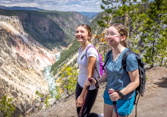 Two girls using hiking poles, walking on a dirt trail and large mountains in the distance