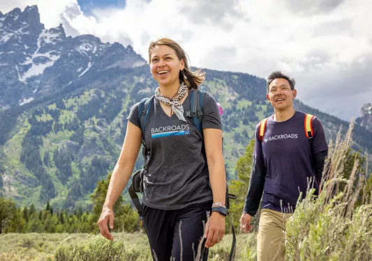 Man and woman smiling while walking through a grassy valley, with large mountains in the background