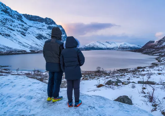 Two people wearing thick jackets looking out to a lake and snowy mountains