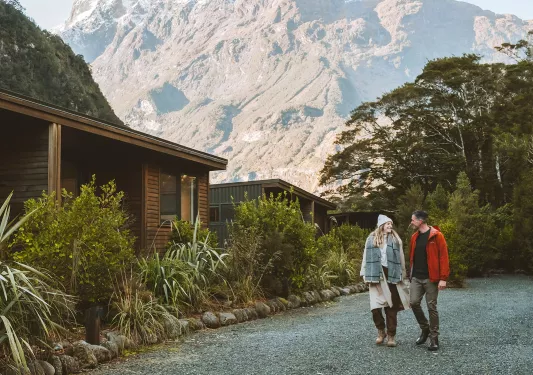 Man and woman walking along a cabin, with large mountains in the background