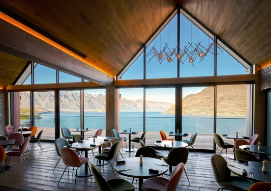 Indoor dining hall with large glass walls, looking out to tall mountains and a lake