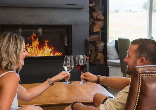 Man and woman smiling in front of a fireplace, holding up glasses of wine