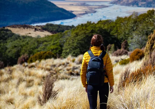 Woman in a yellow jacket walking on a gravel trail, surrounded by tall weeds