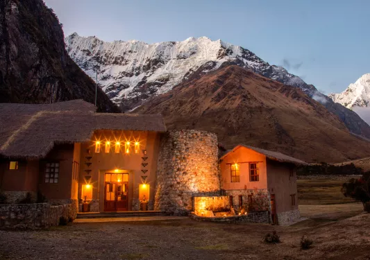 Stone building with straw roofing, with illuminated orange lights and snow-capped mountains in the backgroun