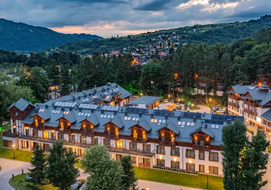 Exterior view of illuminated hotel building with wooden balconies and a forest in the background
