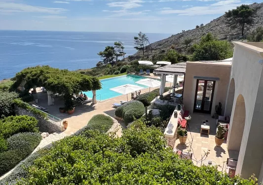 Outdoor pool and beige hotel building, surrounded by trees and views of the ocean in the distance