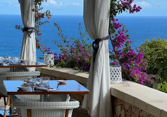Outdoor dining area with wooden tables and woven white chairs, with the ocean in the background