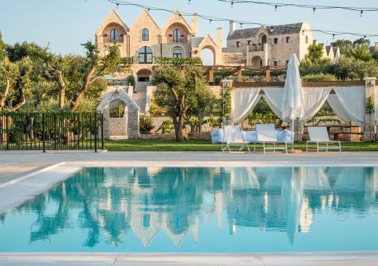 Outdoor pool, surrounded by white chairs and umbrellas, with large stone buildings in the distance