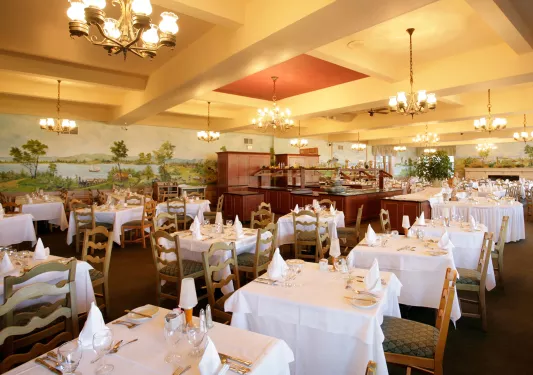 Restaurant dining hall with cushioned chairs and white cloths over dining tables, with a large mural along the wall