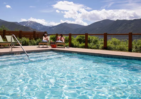 Two women sitting in front of an outdoor pool, with large mountains in the distance