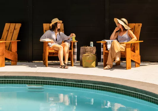 Two women sitting in front of an outdoor pool, laughing and holding cans of drinks