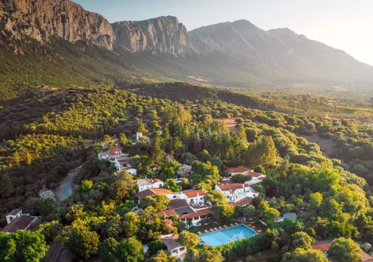 Sky view of a white and red hotel complex in the middle of a forest, with tall mountains in the background
