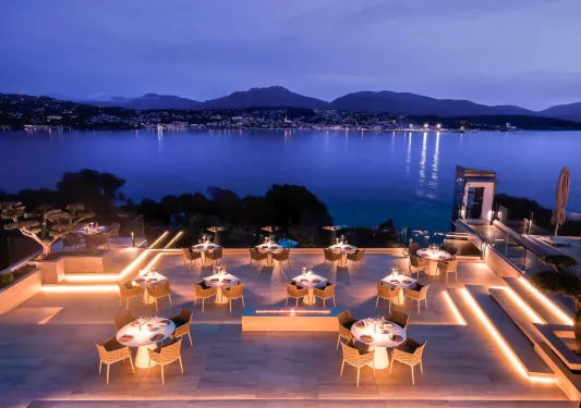 Illuminated outdoor patio with woven chairs and white, circular tables, looking out towards a large lake