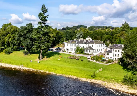 Exterior view of white and gray hotel building, with a grass field and river in front