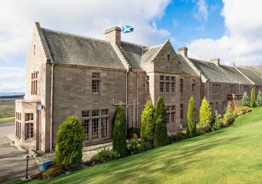 Rustic, stone building with a blue flag on top, with a grass field in the back