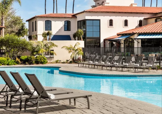 Outdoor pool surrounded by reclining chairs, with large trees and a white building in the background