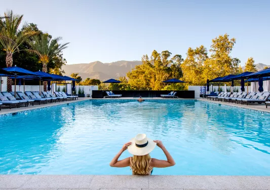 Woman wearing a white hat, standing in an outdoor pool looking towards tall mountains