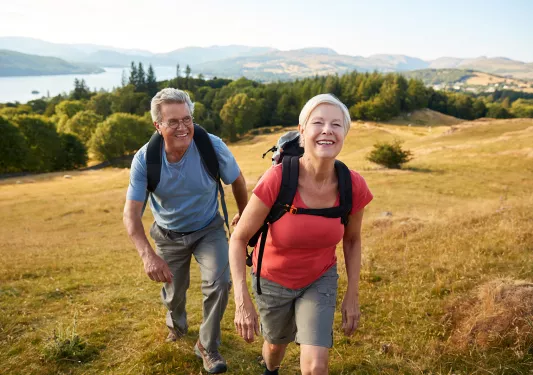 Man and woman ascending a dried, grassy hill with trees in the background