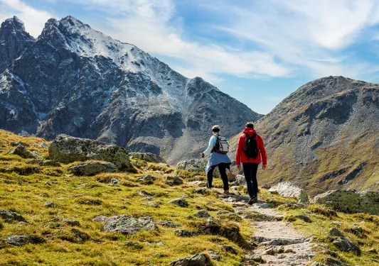 Man and woman ascending a rocky, dirt trail with large mountains ahead