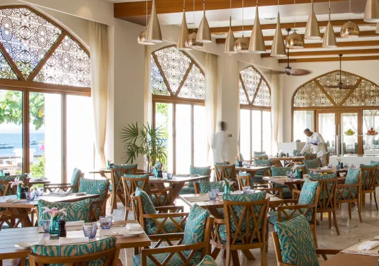 Restaurant dining hall with cushioned chairs surrounding dining tables, with large windows to the left