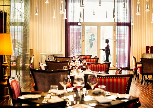 Indoor dining hall with red and tan cushioned chairs, with wine glasses on each table