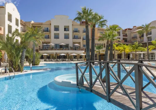 Beige hotel building with an outdoor pool in front, with large palm trees
