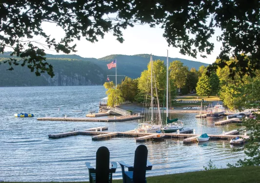 Two small, wooden chairs on a field of grass, overlooking a lake docked with boats