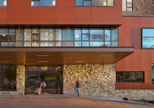 Exterior view of red building with exposed brick, and people walking in with suitcases