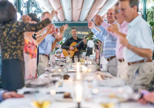 Group of people at a dining table, dancing while a man with a guitar is singing