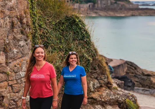 Two women walking along a stone back with hills and the ocean in the background