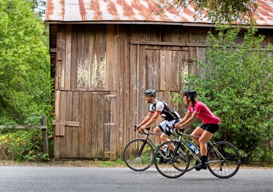 Man and woman wearing biking gear, riding bikes in front of a wooden barn on a road