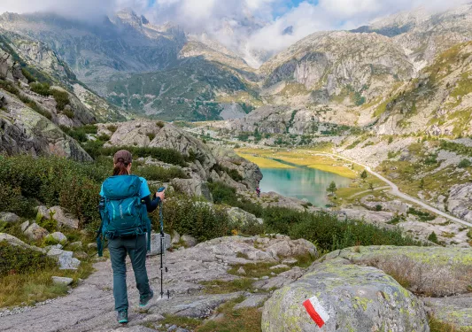 Woman with a blue backpack descending down a rocky hiking trail with mountains in the distance
